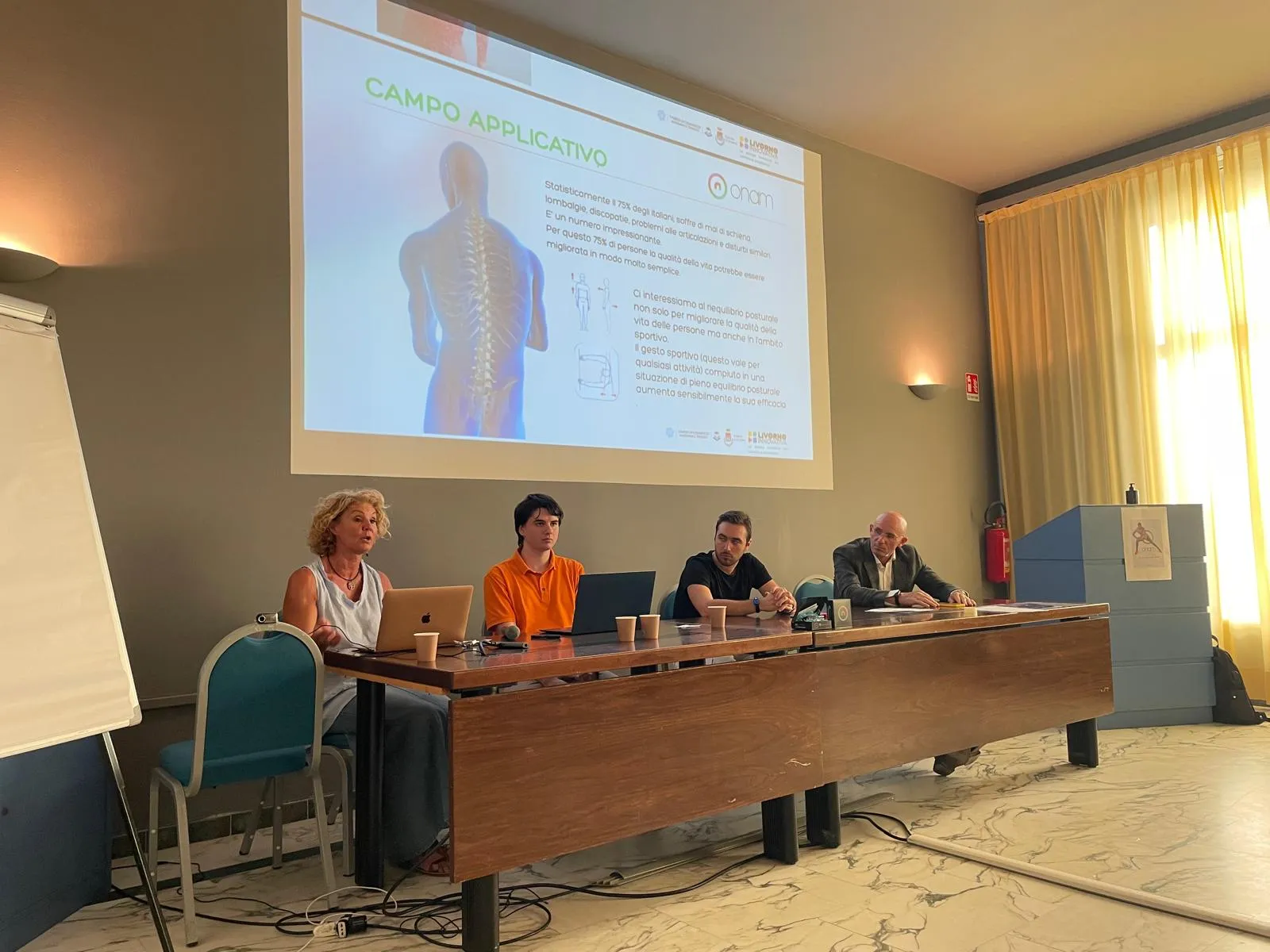 Quattro persone del team ONAM durante una presentazione pubblica, con alle spalle una slide sul campo applicativo del dispositivo